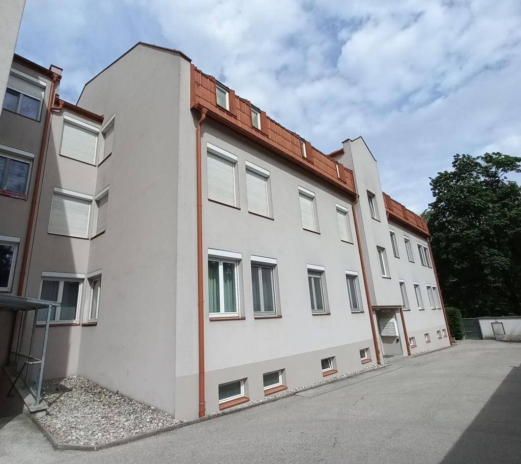 Mehrstöckiges Wohngebäude mit heller Fassade, roten Dachziegeln und vielen Fenstern mit Rollläden.