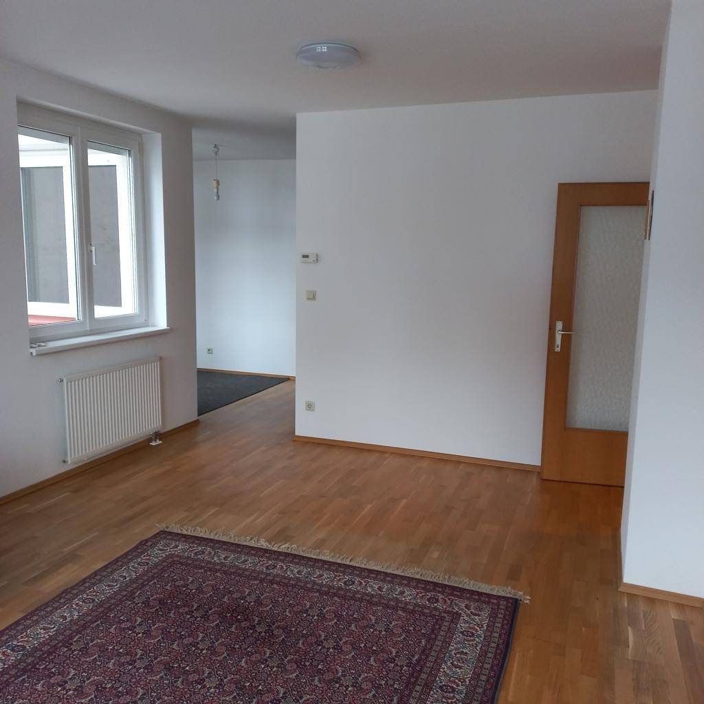 Heller Wohnbereich mit Holzfußboden und Teppich, der offen zu einem weiteren Raum führt.
