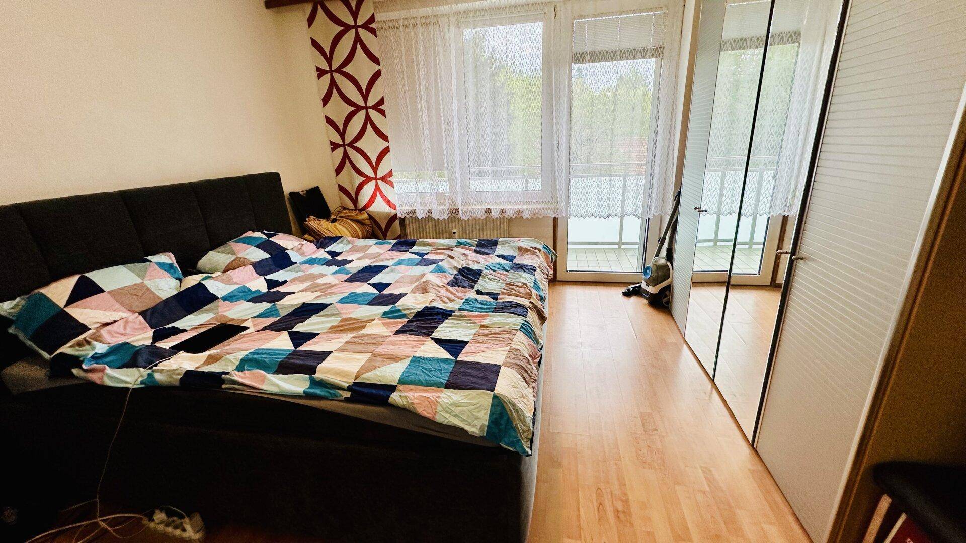 Schlafzimmer mit Doppelbett, Holzboden und Spiegelschrank, hell durch große Fenster.
