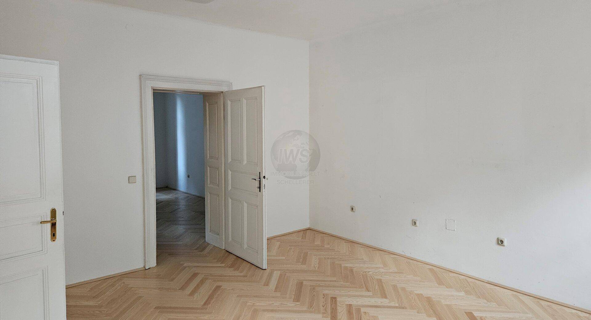 Geräumiges Zimmer mit Fischgrätparkett, weißen Wänden und offener Tür zu einem weiteren Raum.