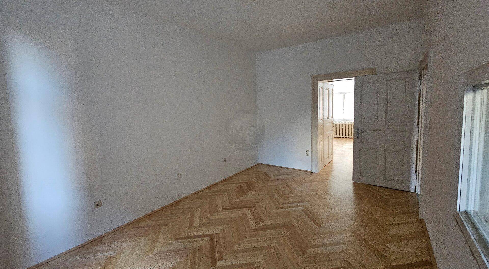 Heller Raum mit Fischgrätparkett und offener Tür, die den Blick in ein weiteres Zimmer freigibt.
