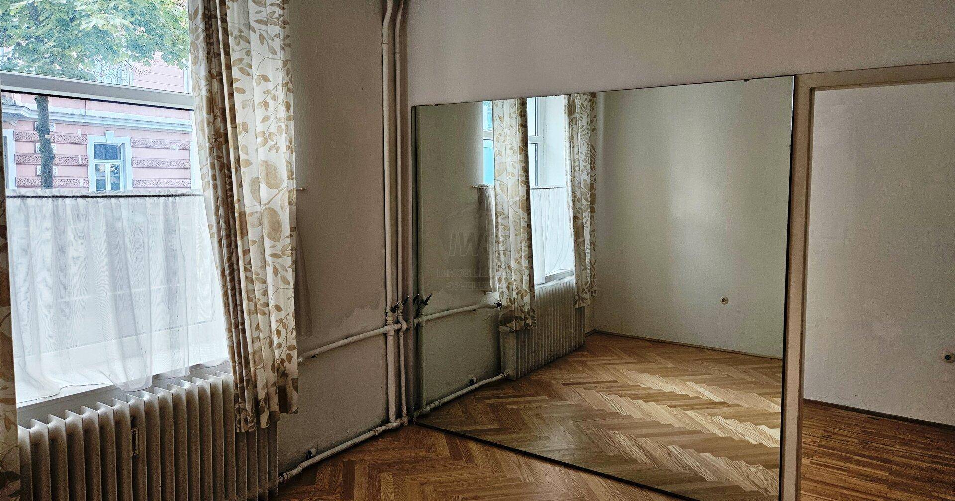 Zimmer mit Fischgrätparkett, großem Spiegel und Fenster mit Vorhängen, das viel Licht hereinlässt.