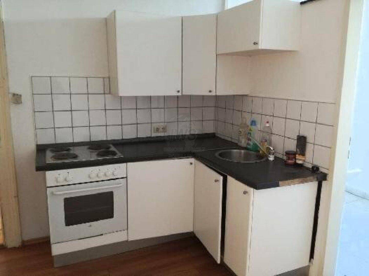 Kleine Eckküche mit weißen Schränken, Spüle, Elektroherd und weißem Fliesenspiegel.