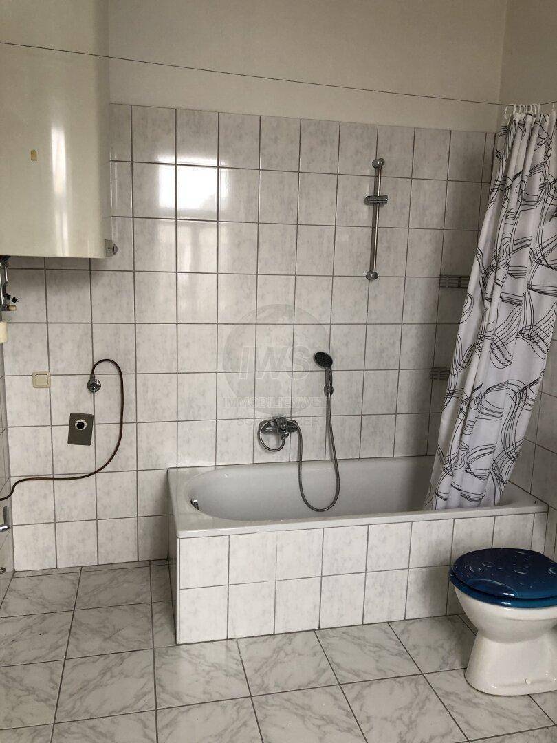 Badezimmer mit weißen Fliesen, Badewanne mit Duschfunktion und WC mit blauem Sitz.