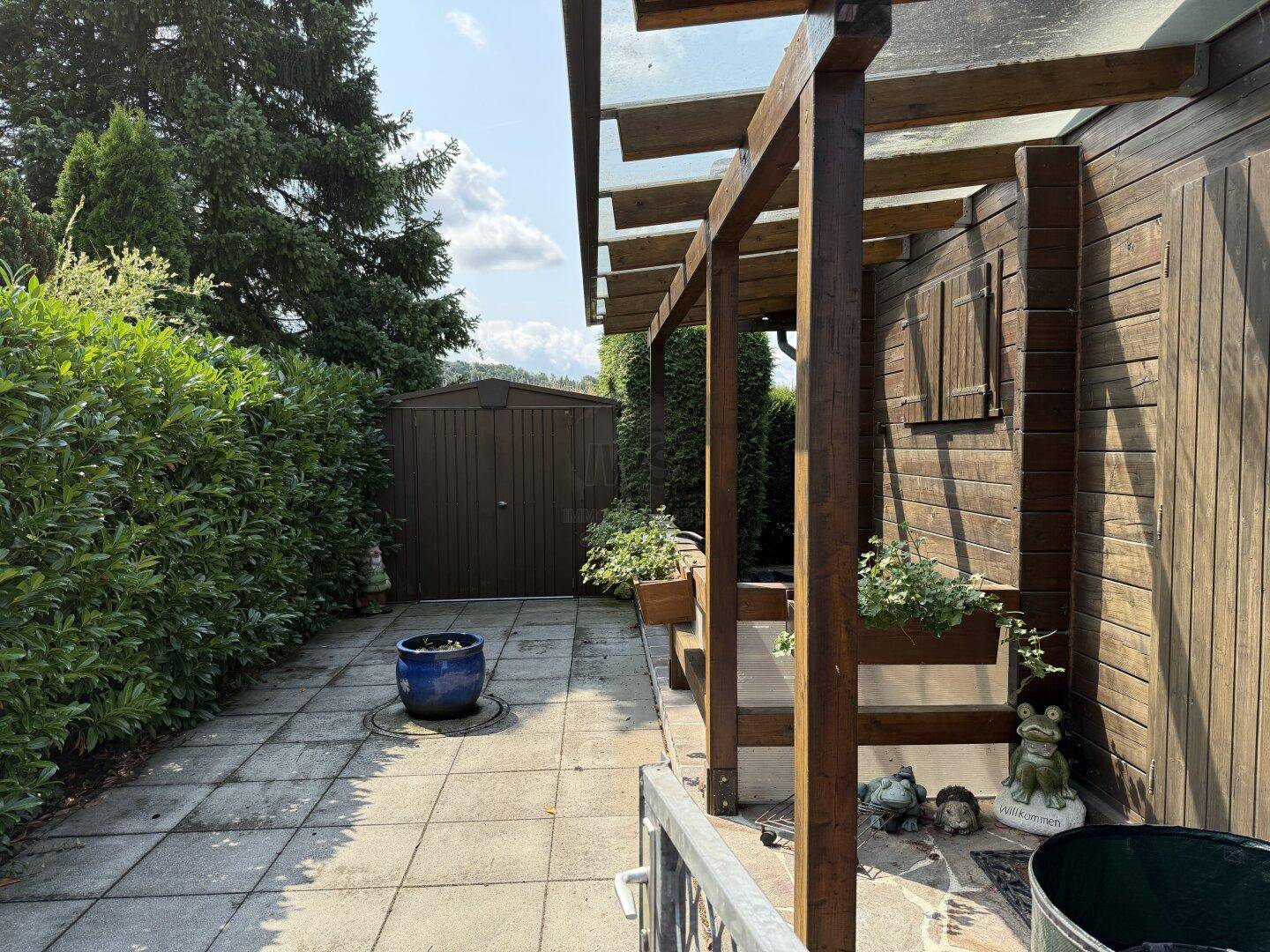 Gepflasterter Weg neben dem Holzhaus, führt zu einem Gartenschuppen unter einer Holzpergola.