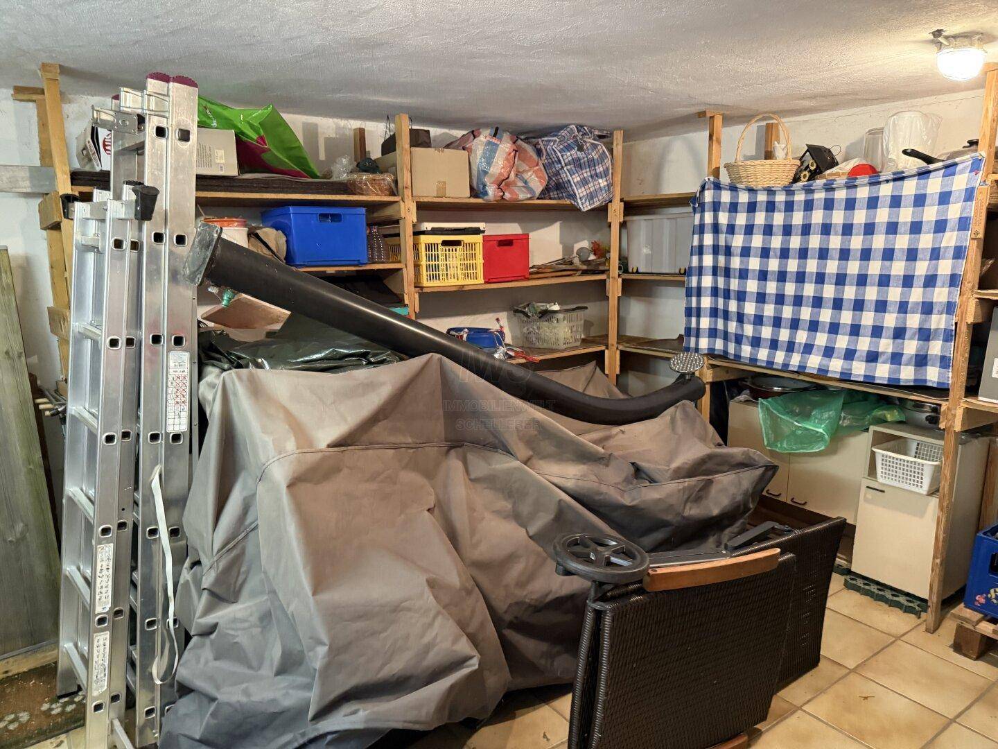 Unaufgeräumter Abstellraum im Keller mit Regalen voller Gegenstände und einer Leiter.