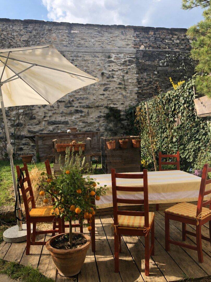 Gemütlicher Essbereich im Garten auf einer Holzterrasse mit Sonnenschirm und Steinmauer.