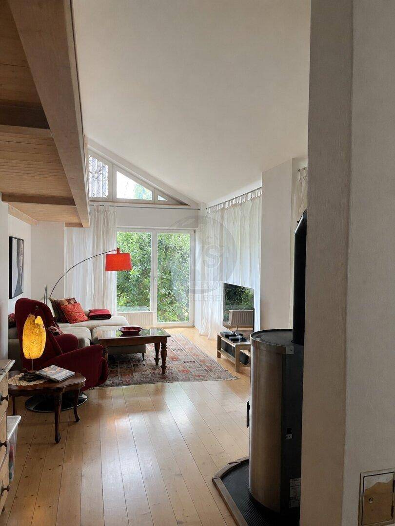 Helles Wohnzimmer mit Kaminofen, Holzboden und großen Fenstern mit Blick ins Grüne.