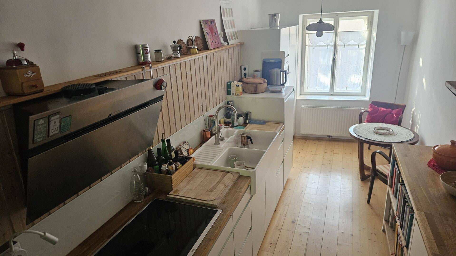 Helle Küche mit Holzboden, weißen Schränken und einem kleinen Essbereich am Fenster.