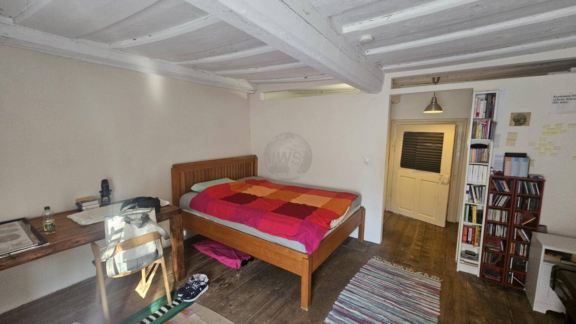 Schlafzimmer mit Holzbalkendecke, Holzboden und Bücherregalen, gemütlich eingerichtet.
