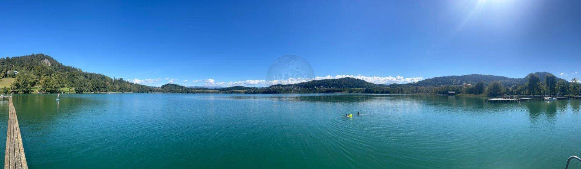 Panoramablick auf einen klaren See mit grünen Hügeln und Bergen unter strahlend blauem Himmel.