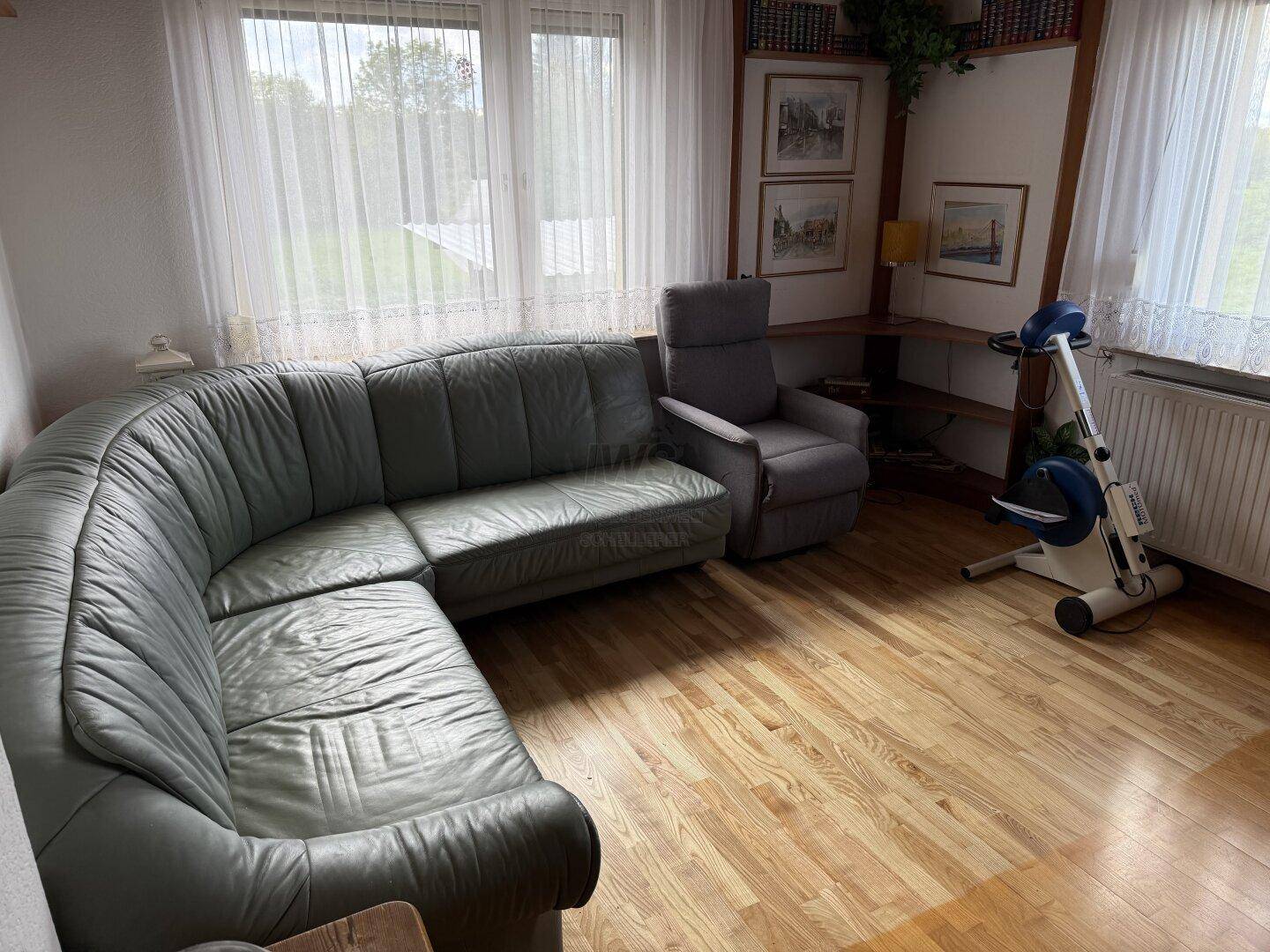 Gemütliches Wohnzimmer mit grauem Ledersofa, Holzfußboden und großen Fenstern.