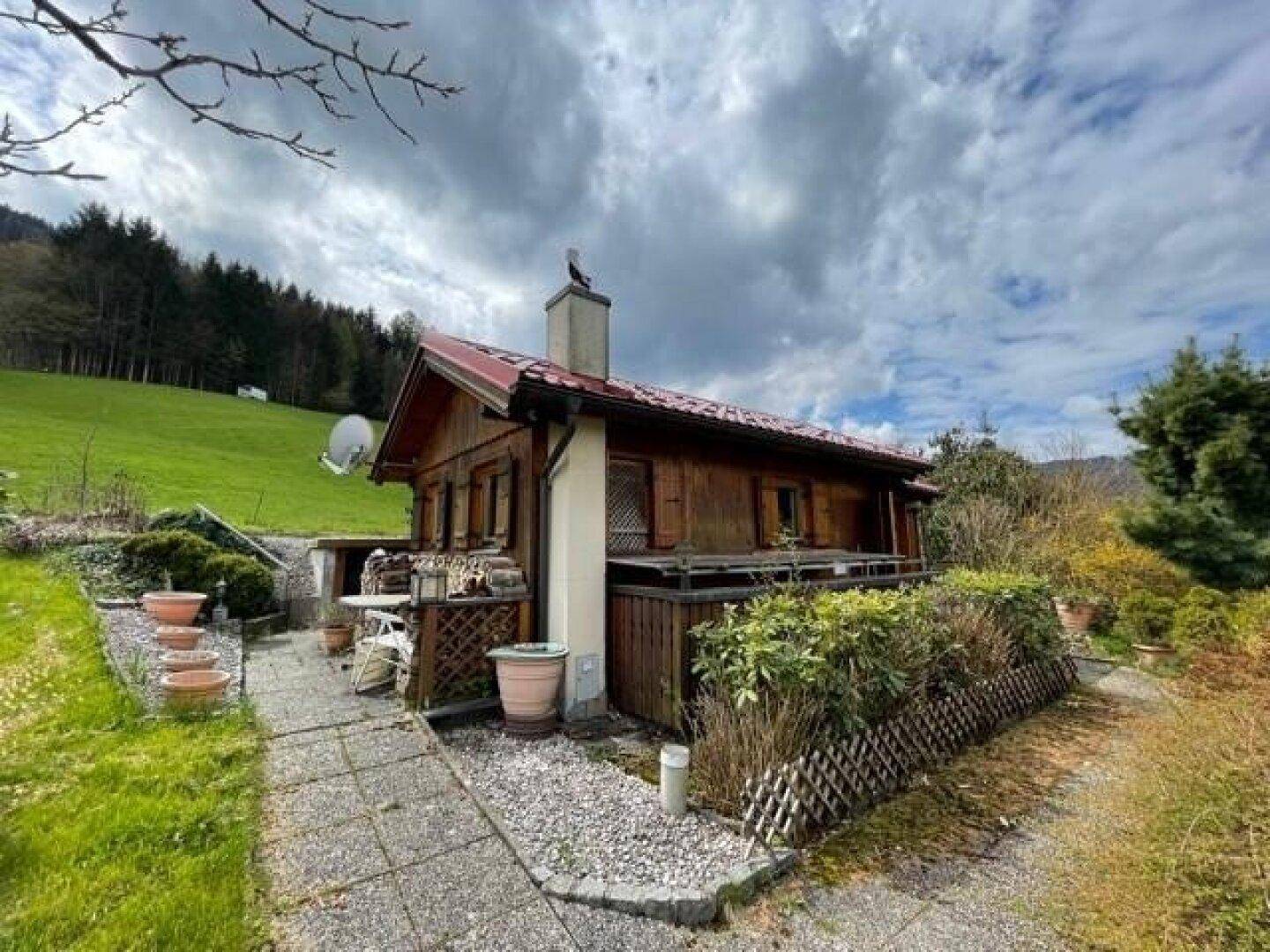 Rustikales Holzhaus mit rotem Dach, kleiner Terrasse und gepflegtem Gartenbereich vor grüner Hügellandschaft.