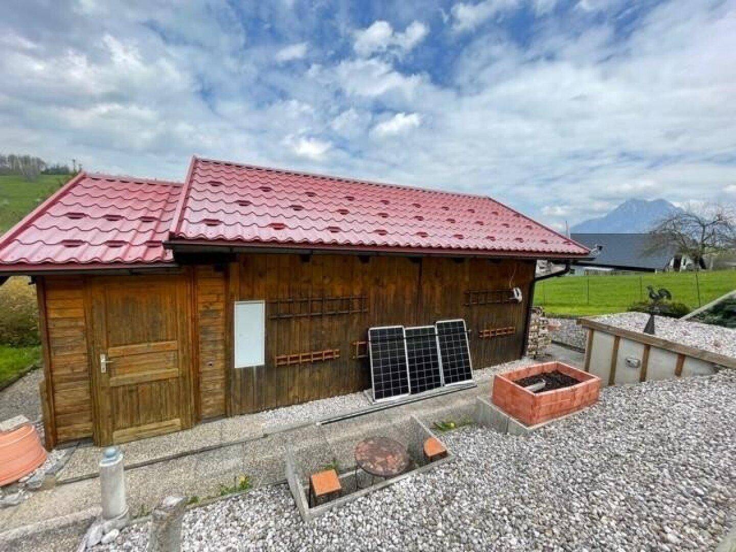 Seitenansicht des Holzhauses mit rotem Dach, Kiesfläche und Solarmodulen, die an der Wand lehnen.