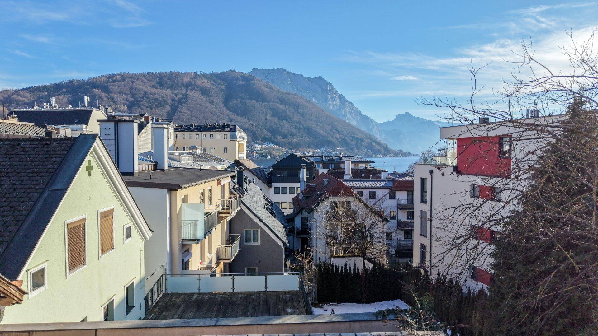 Panoramablick über die Dächer einer Stadt mit einem See und majestätischen Bergen im Hintergrund unter blauem Himmel.