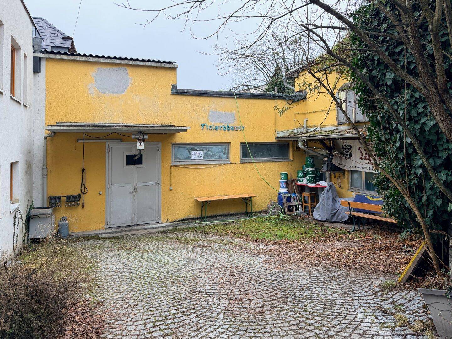 Außenansicht eines gelben Gebäudes mit gepflastertem Hof und Grünflächen.