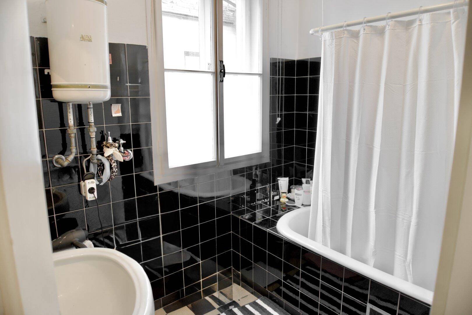 Badezimmer mit schwarzen Fliesen, Badewanne mit Duschvorhang und Fenster für Tageslicht.