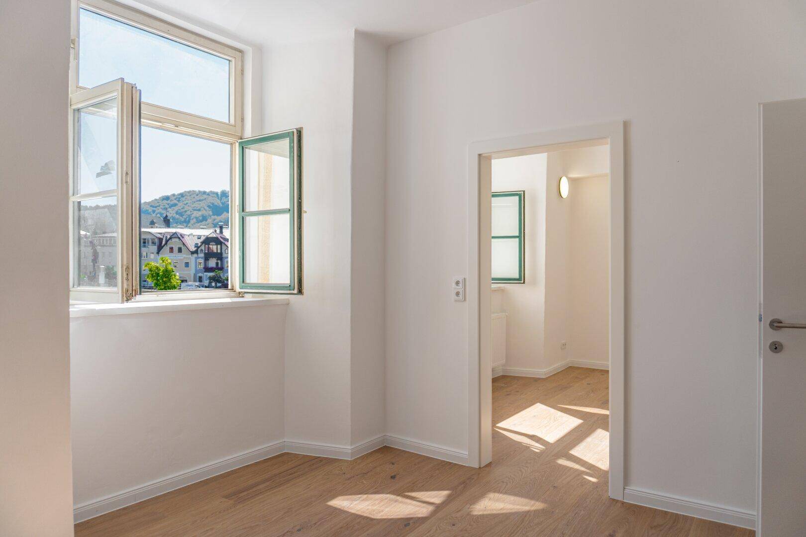 Helles Zimmer mit Holzboden und Fensterblick auf die grüne Umgebung und die Stadt.