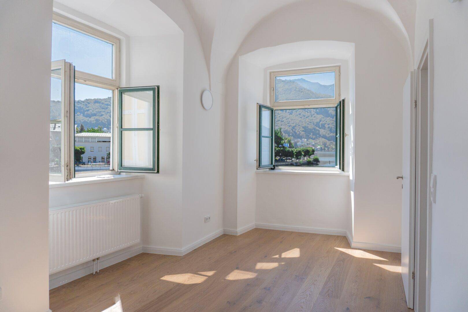 Geräumiges Zimmer mit zwei Fenstern, Holzboden und Blick auf die grüne Umgebung.