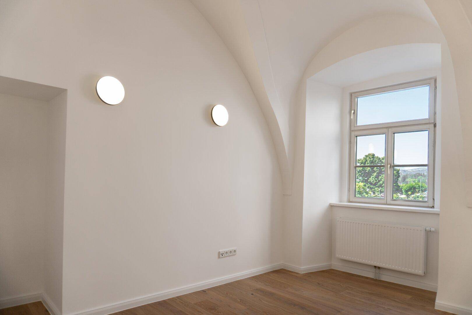 Helles Zimmer mit Gewölbedecke, Holzboden und Fensterblick auf die grüne Umgebung.
