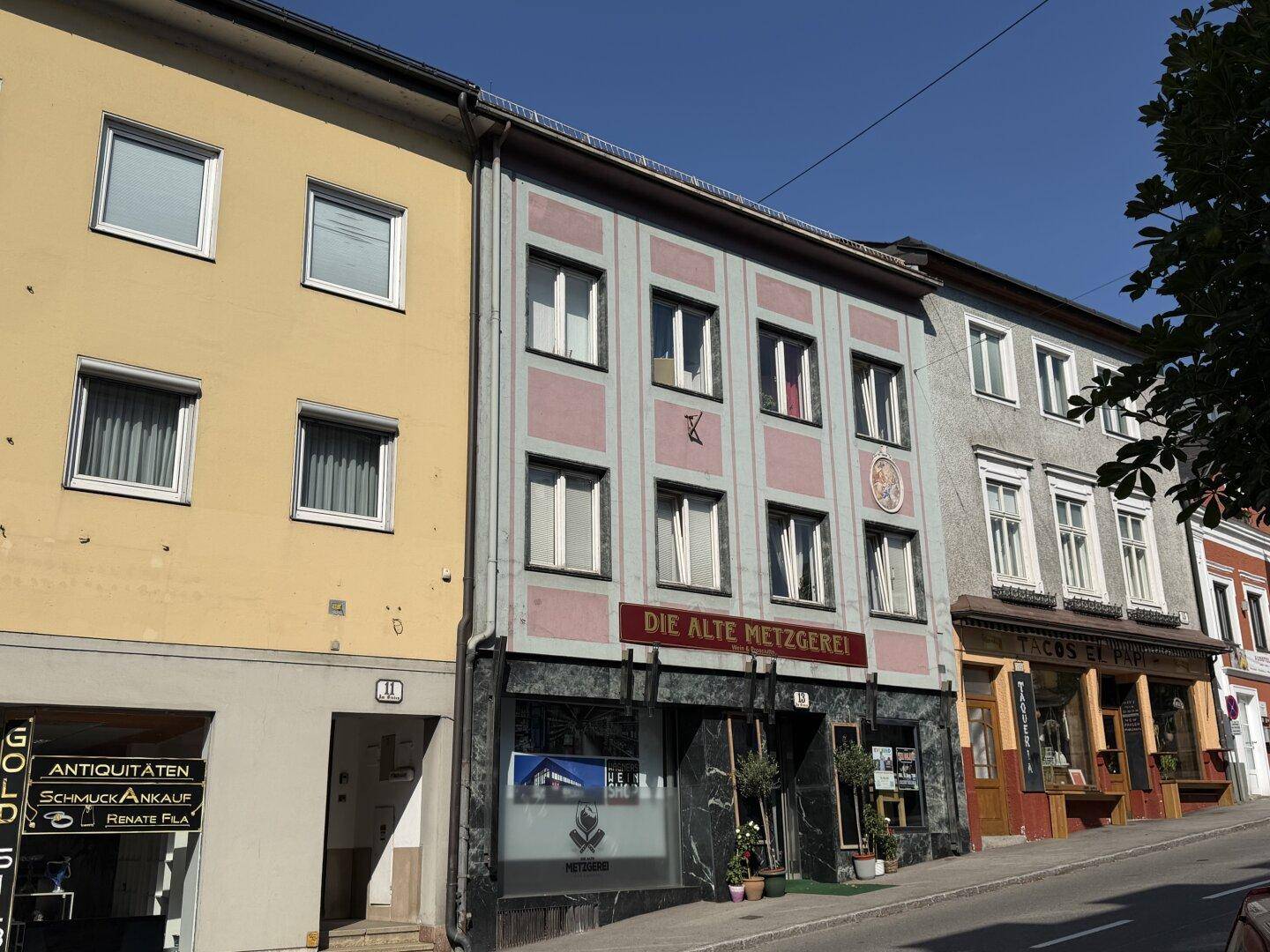 Historische Gebäudezeile mit Geschäften im Erdgeschoss und Wohnungen darüber unter blauem Himmel.