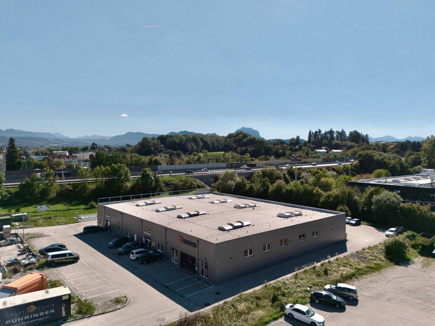 Moderne Gewerbehalle mit Parkplätzen und bergiger Landschaft im Hintergrund unter blauem Himmel.