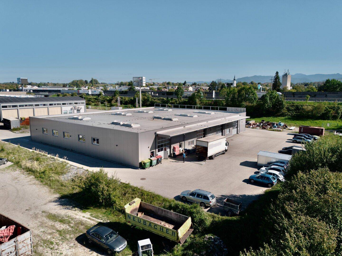 Gewerbegebäude mit Laderampe und großem Parkplatz, umgeben von Bäumen und einer weiten Landschaft.