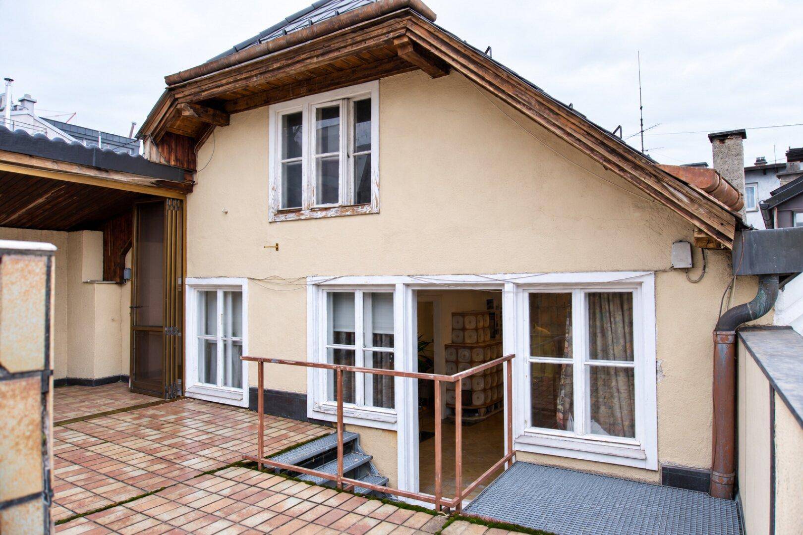 Große geflieste Terrasse an einem Haus mit Holzdachüberstand und mehreren Fenstern.
