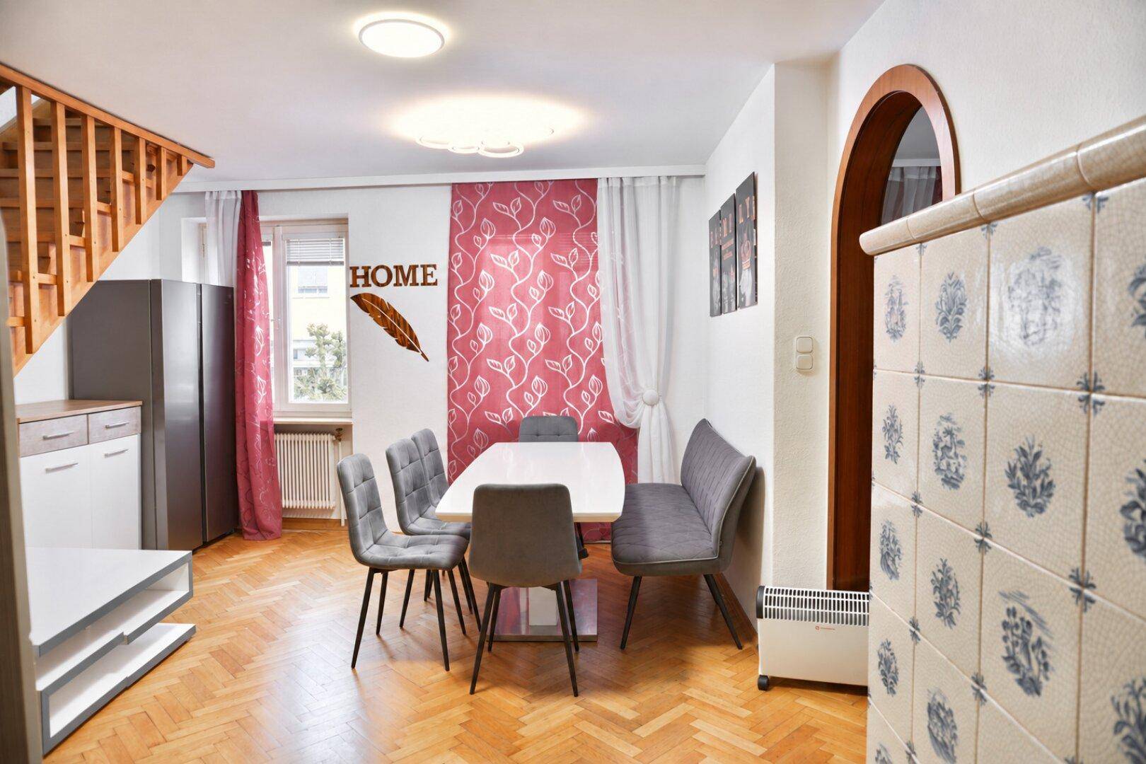Heller Essbereich mit Kachelofen, Holztreppe, modernem Esstisch und roten Vorhängen.