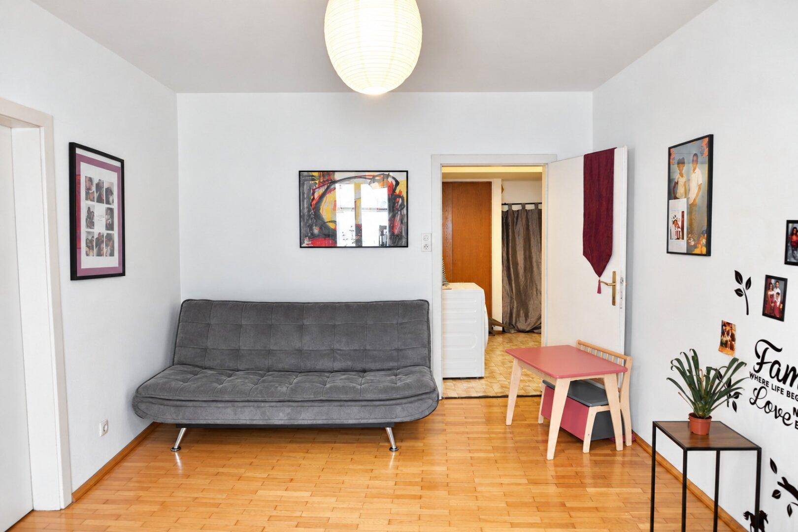 Wohnzimmer mit grauem Sofa, Holzboden und Zugang zu einem Nebenraum.