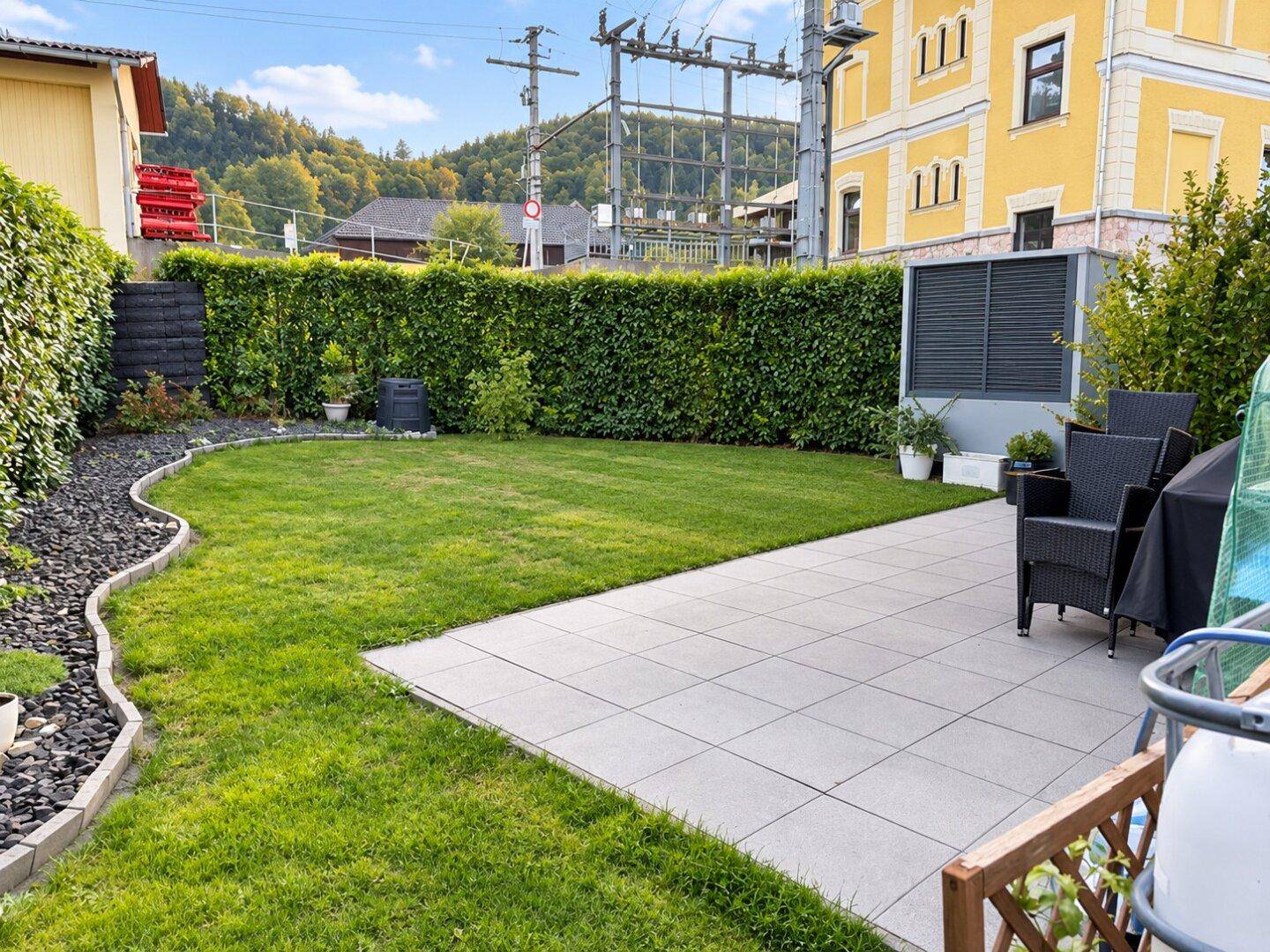 Gepflegter Garten mit Rasenfläche und gepflasterter Terrasse, ausgestattet mit Gartenmöbeln.