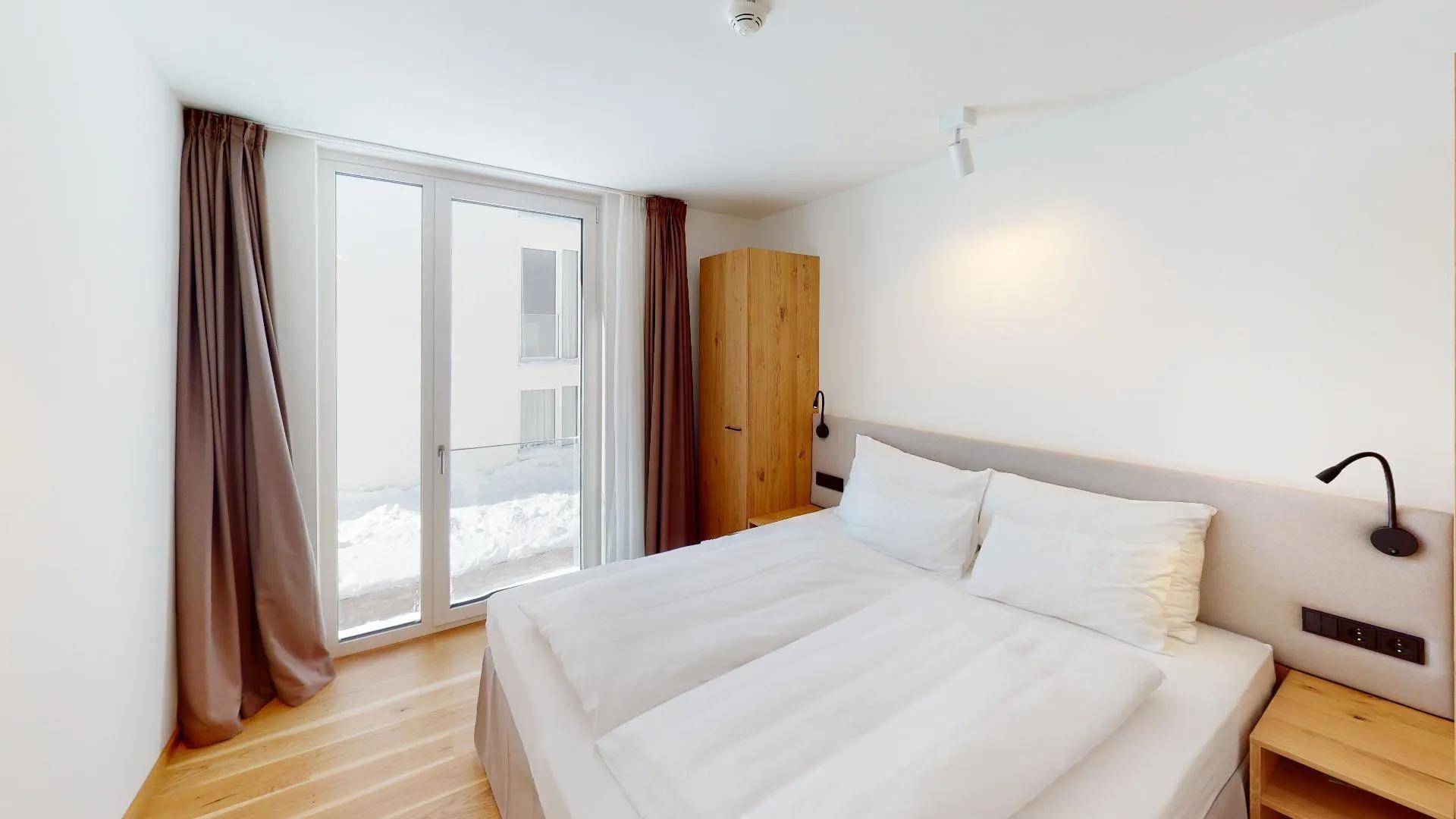 Helles Schlafzimmer mit Doppelbett, Holzschrank und Zugang zu einem Balkon oder einer Terrasse.