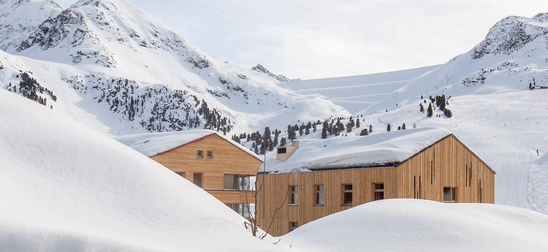 Zwei moderne Holzgebäude eingebettet in eine weite, schneebedeckte Berglandschaft.