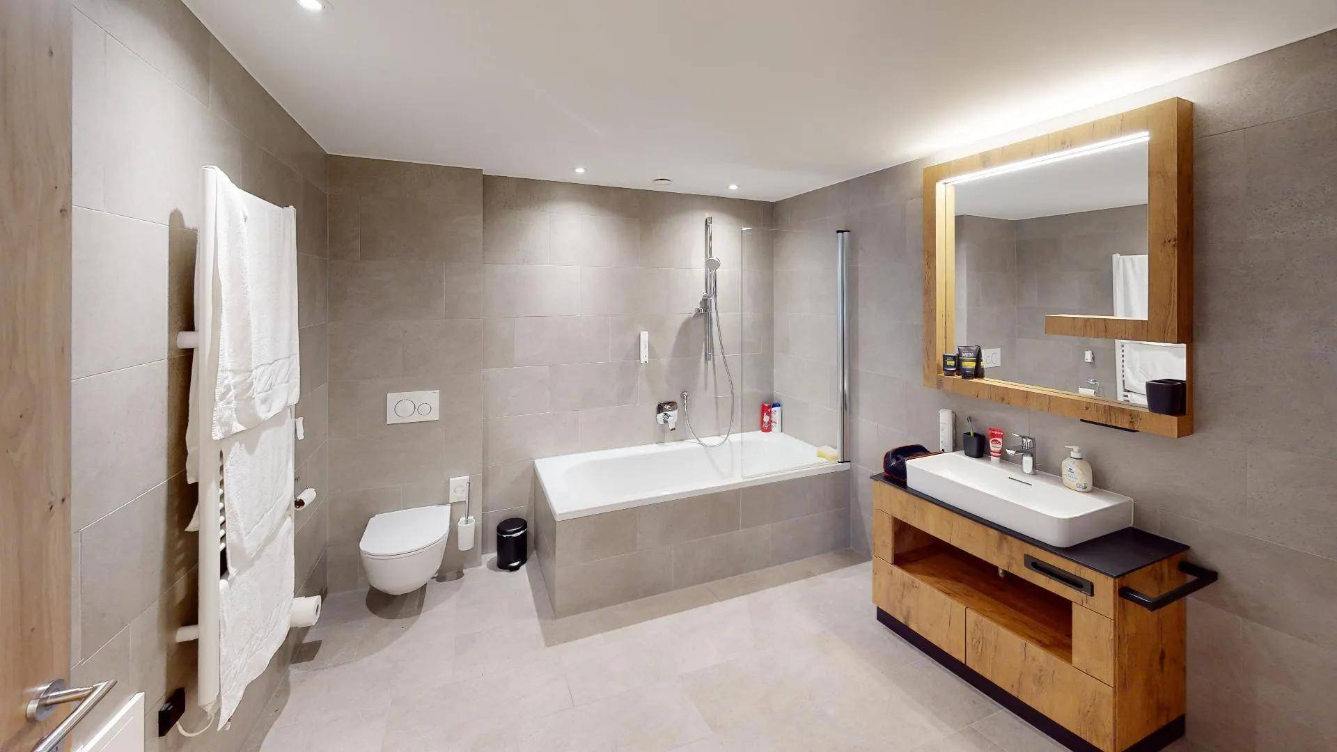 Modernes Badezimmer mit großen Fliesen, Badewanne und stilvollem Holzwaschtisch mit beleuchtetem Spiegel.