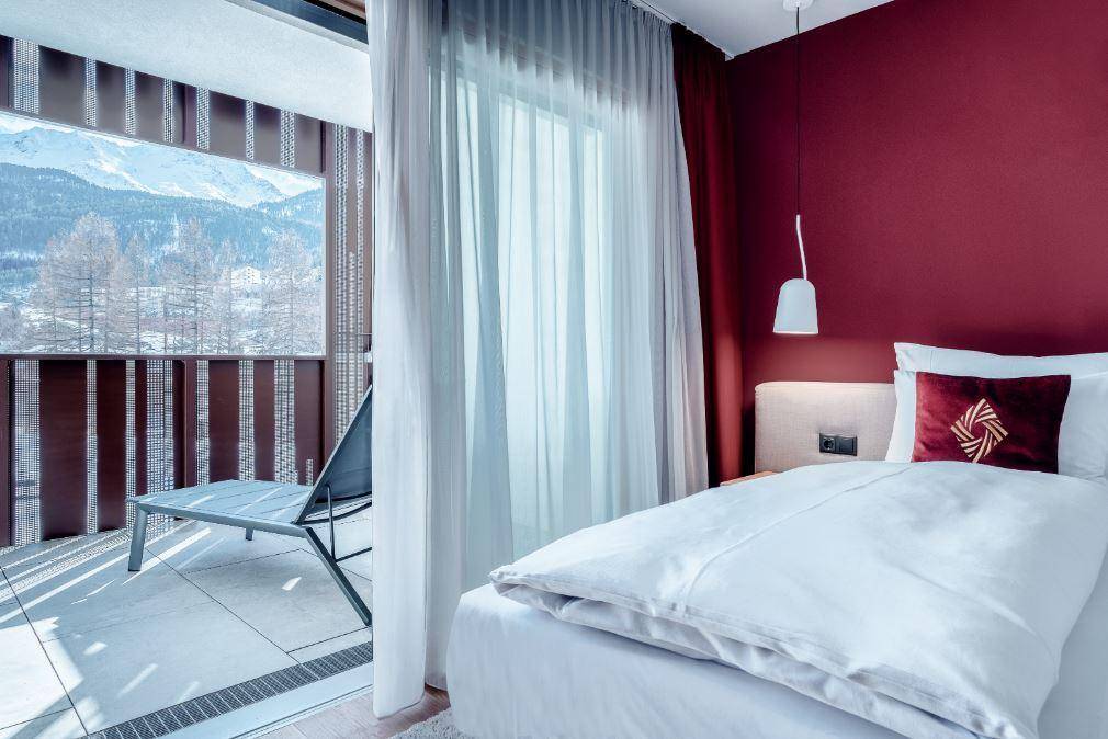 Helles Schlafzimmer mit roter Akzentwand, bequemen Bett und direktem Zugang zum Balkon mit Bergblick.