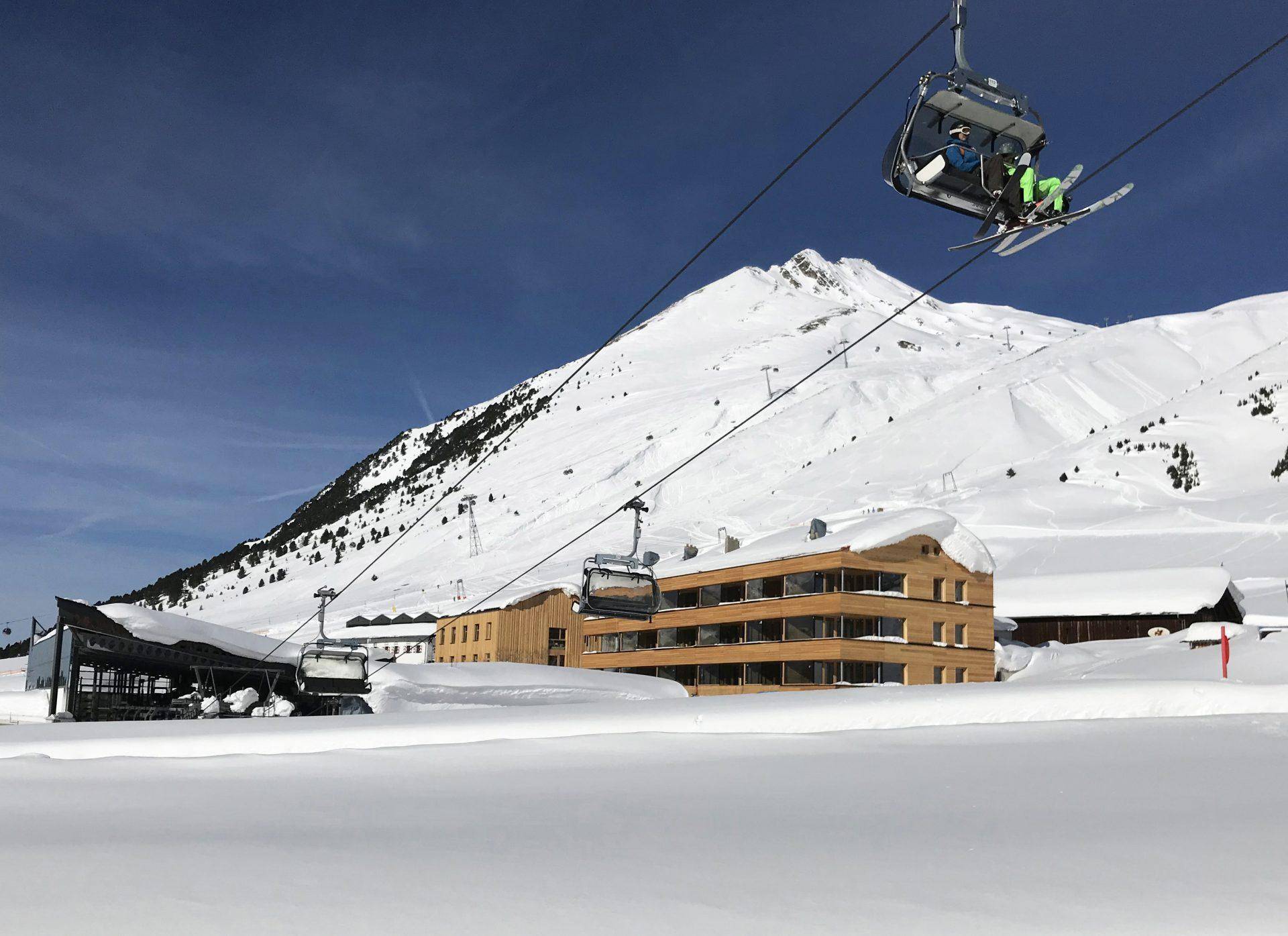 Skigebiet mit Sessellift, modernen Gebäuden und schneebedeckten Bergen unter blauem Himmel.