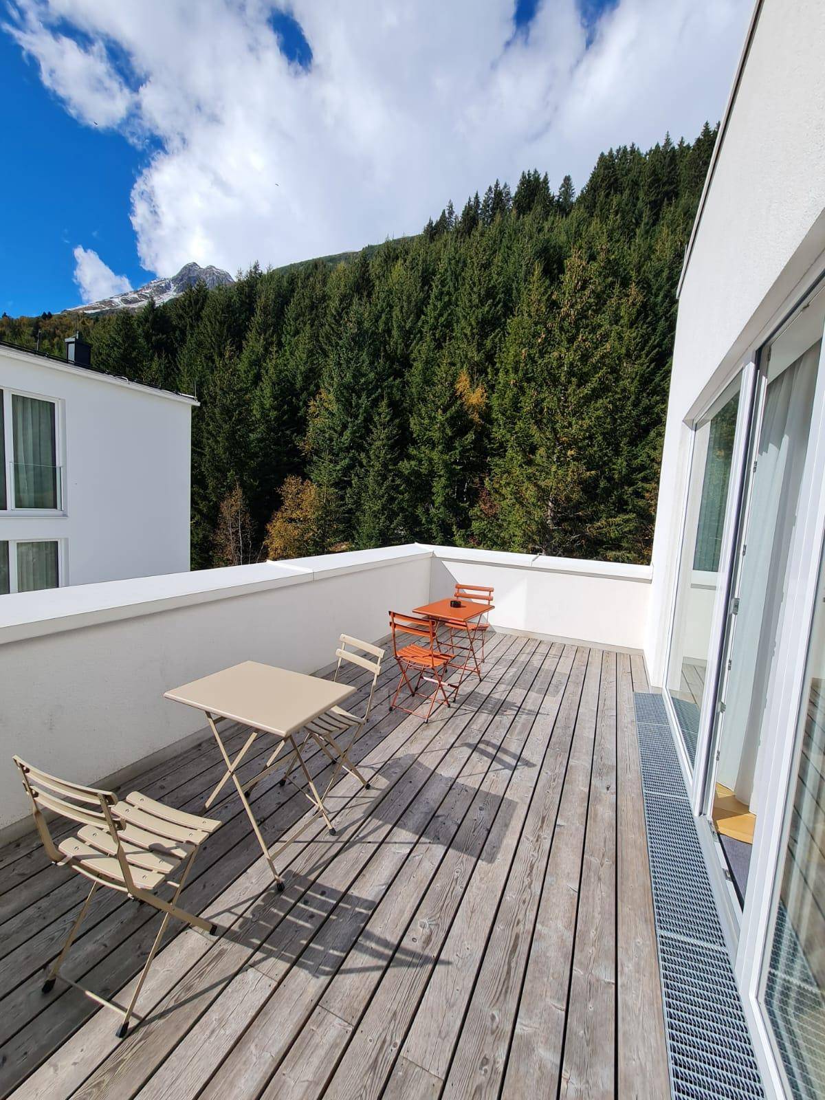 Sonniger Balkon mit Holzboden und einfacher Bestuhlung, umgeben von grünen Bergen.