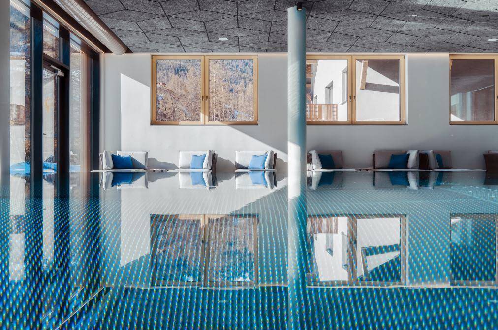 Heller Innenpool mit blauen Fliesen, großen Fenstern und bequemen Liegeflächen am Beckenrand.