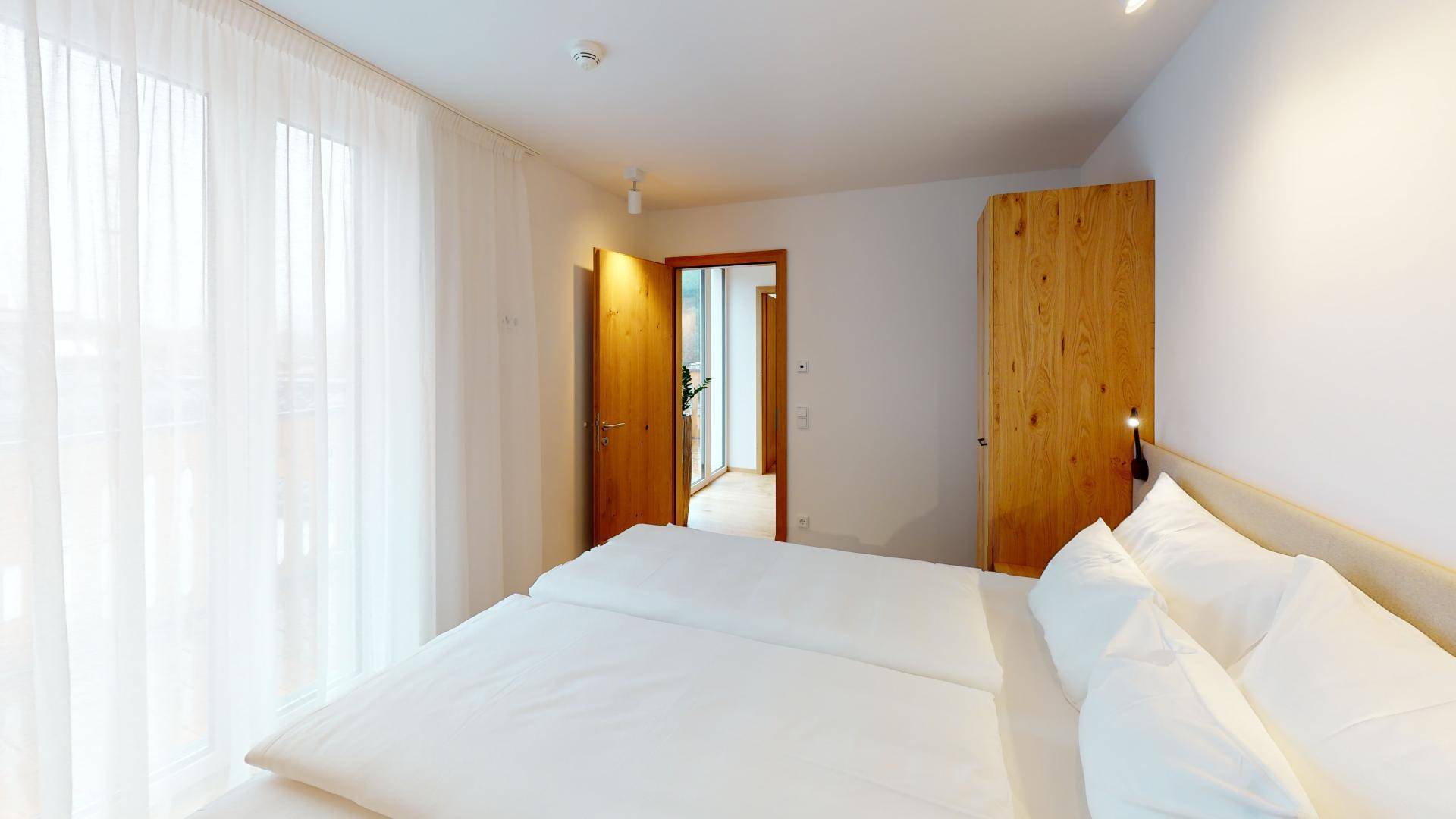 Helles Schlafzimmer mit großem Fenster, transparenten Vorhängen und Zugang zu einem weiteren Raum.