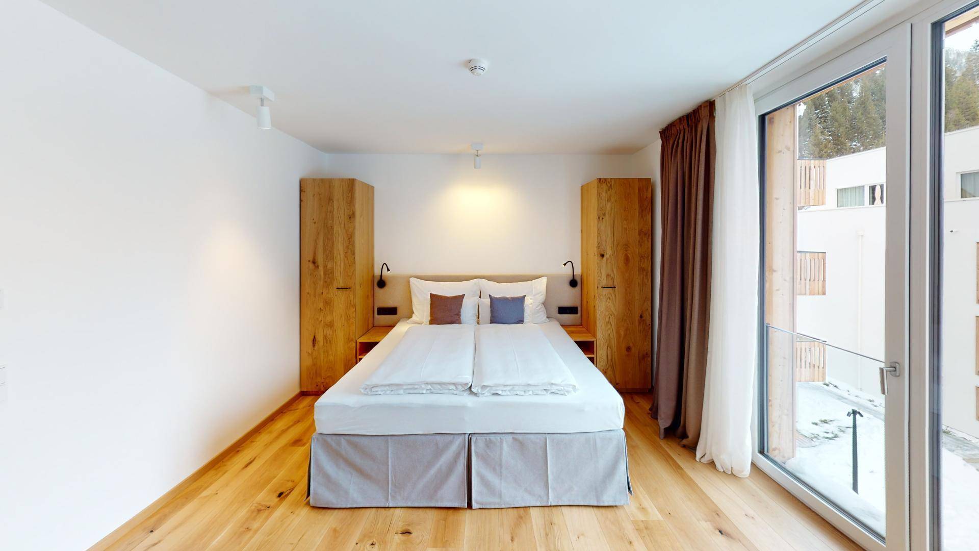 Modernes Schlafzimmer mit Doppelbett, Holzboden und großen Fenstern, die viel Licht hereinlassen.