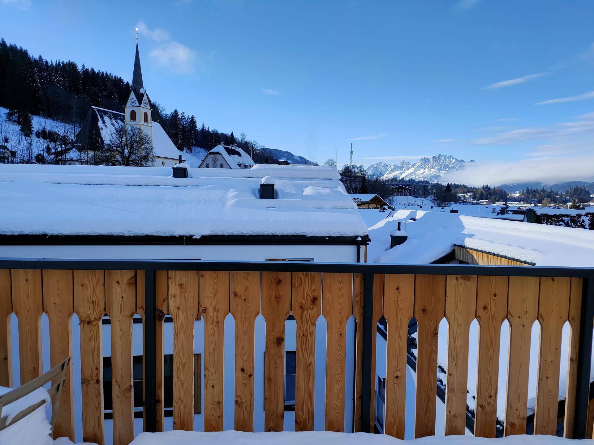 Balkonblick auf ein verschneites Dorf mit einer Kirche und majestätischen Bergen im Hintergrund.