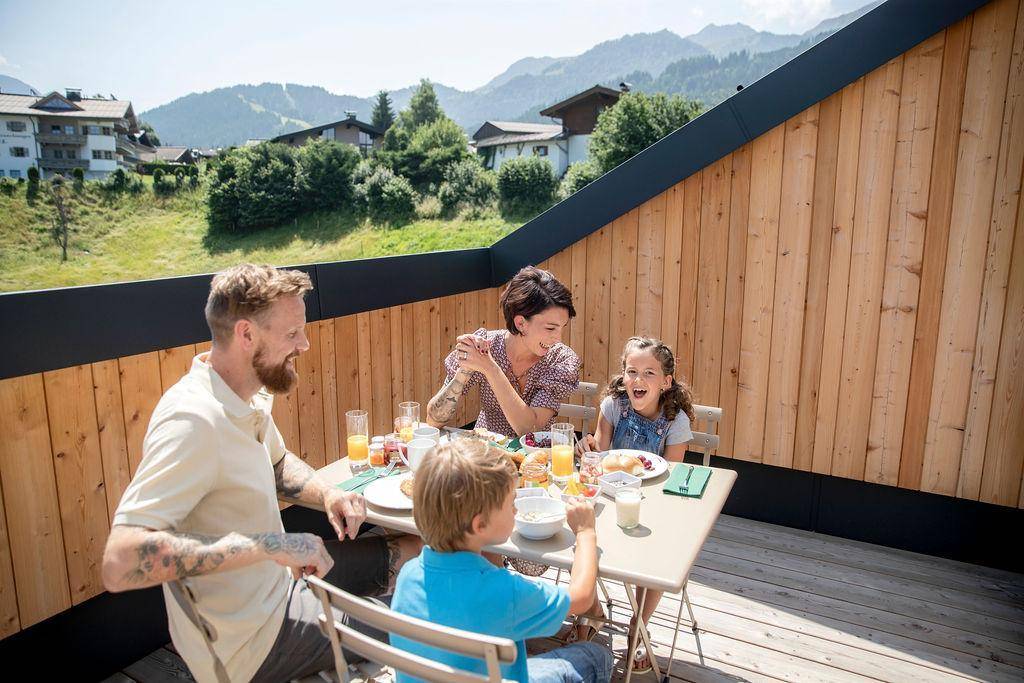 Fröhliche Familie beim Frühstück auf der Dachterrasse mit Panoramablick auf die umliegende Landschaft.