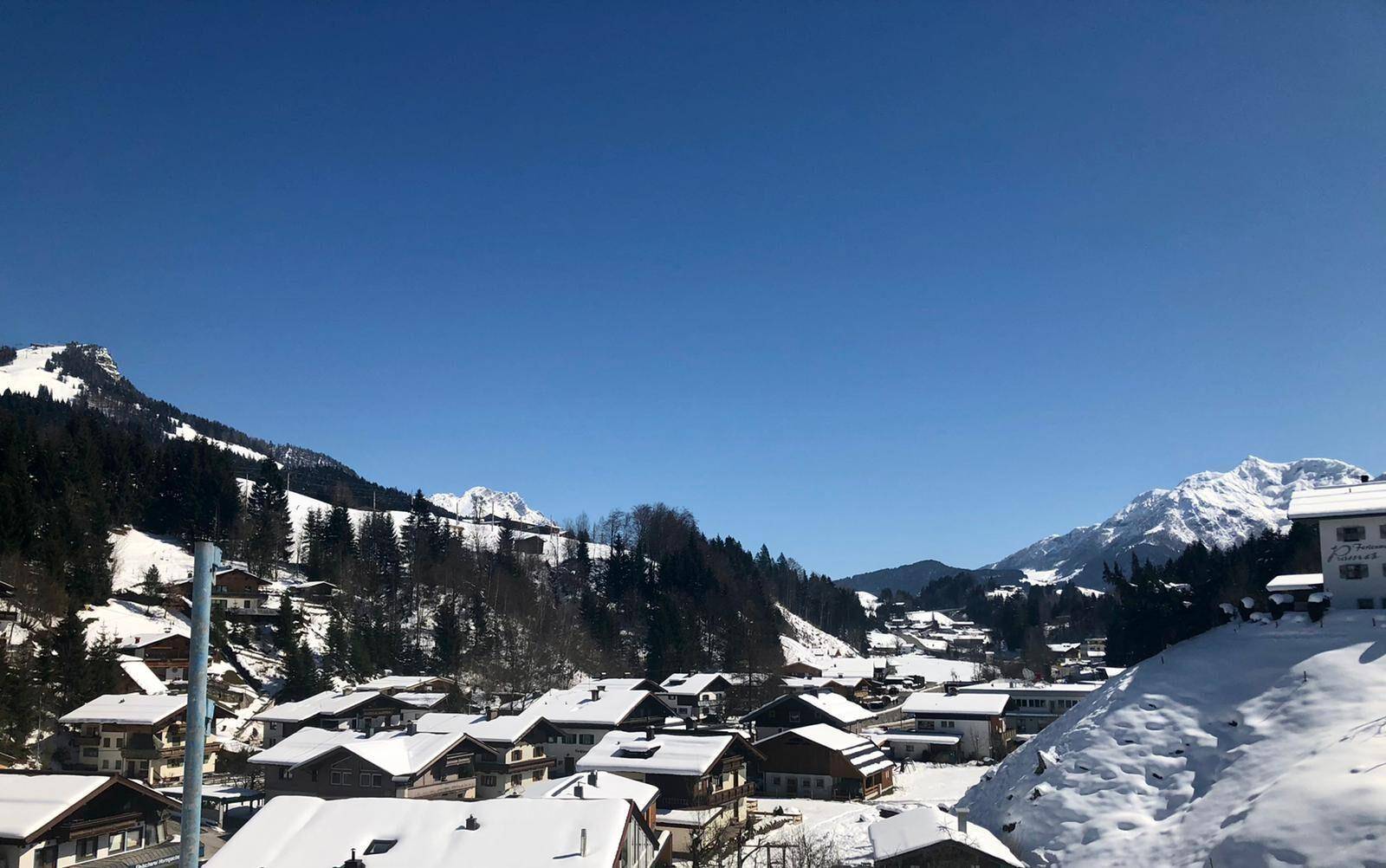 Winterlicher Panoramablick auf ein idyllisches Bergdorf, umgeben von schneebedeckten Bergen und Wäldern.