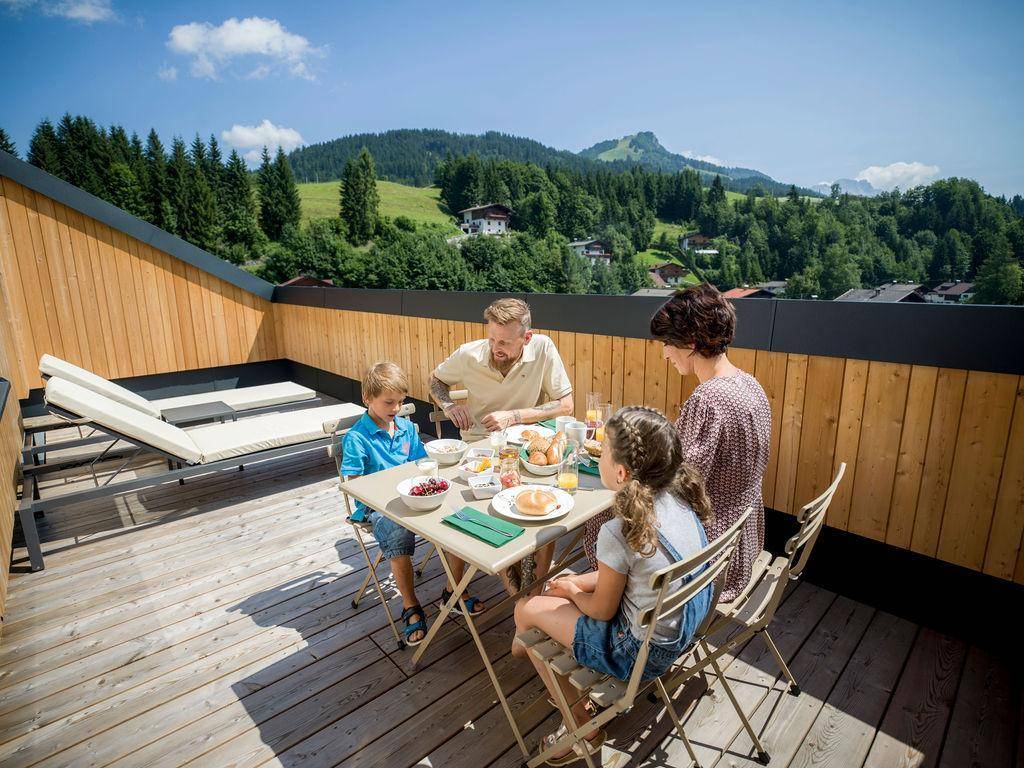 Familie genießt ein Frühstück auf einer sonnigen Dachterrasse mit Holzboden und Bergblick.