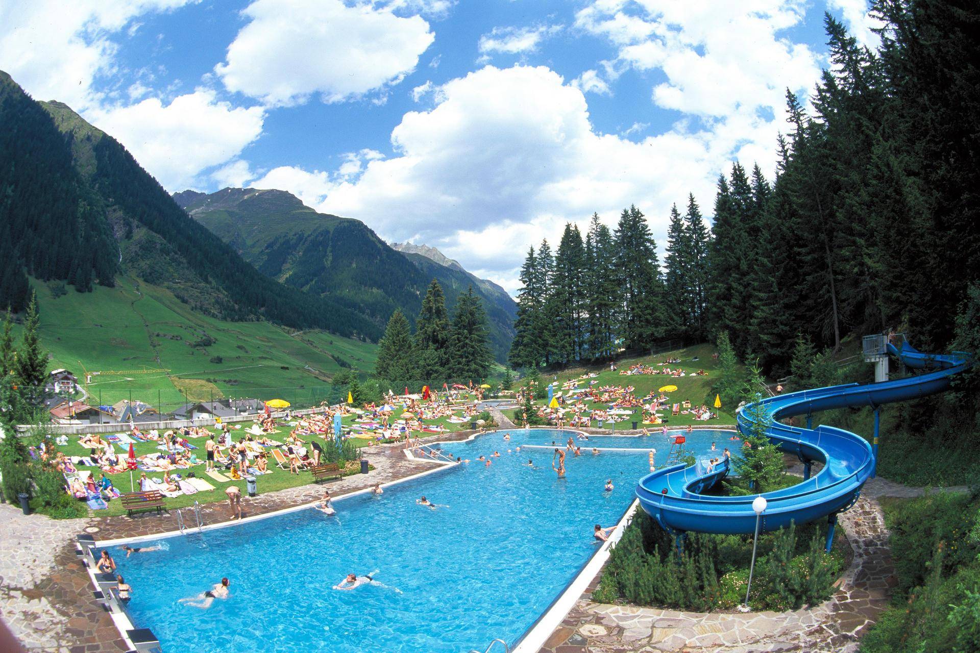 Großes Freibad mit Wasserrutsche, umgeben von grünen Wiesen und Bergen an einem sonnigen Tag.