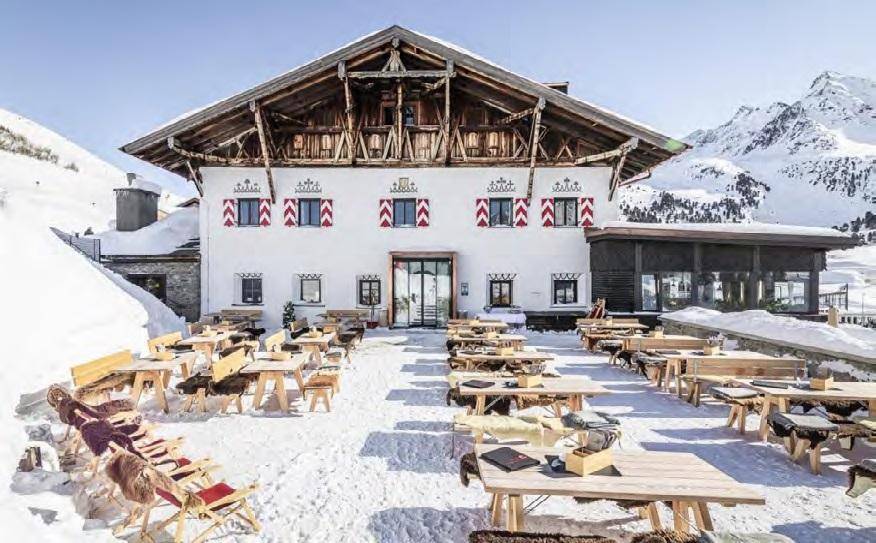 Traditionelles Gebäude mit rustikalen Holztischen und Bänken im Schnee vor Bergkulisse.