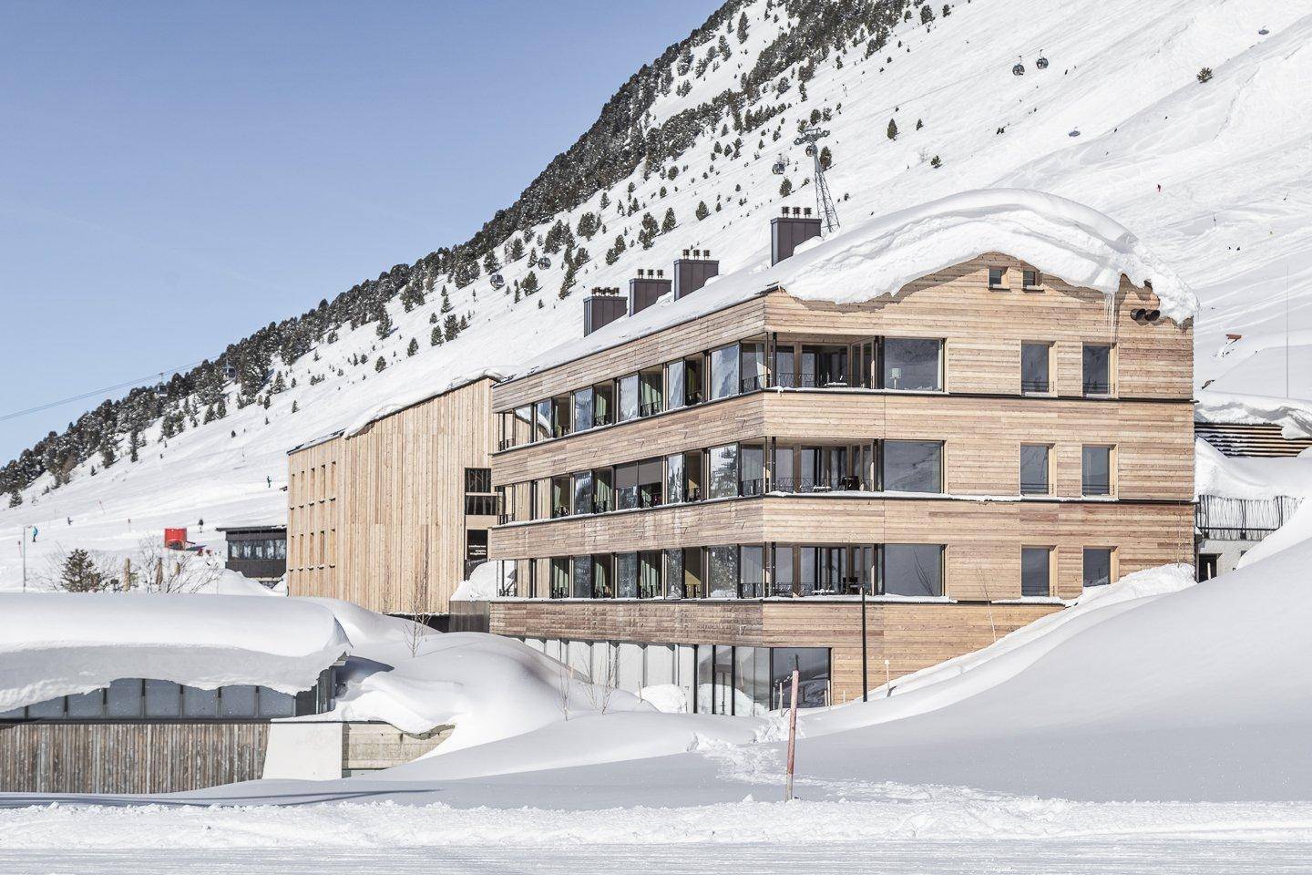 Modernes Holzgebäude in einer verschneiten Berglandschaft mit vielen Fenstern.