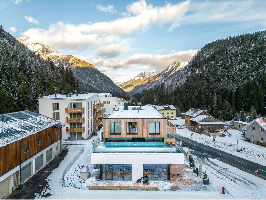 Luftaufnahme eines modernen Gebäudes mit Dachpool, umgeben von verschneiten Bergen und Wäldern.