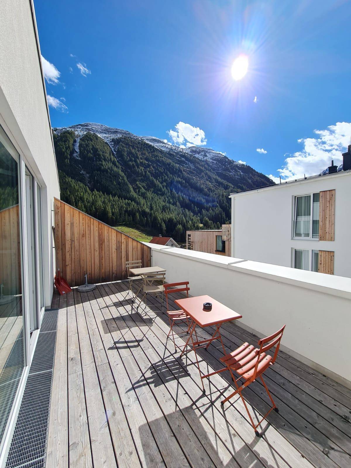 Helle Holzterrasse mit Sitzgelegenheiten und weitem Blick auf die sonnigen Berge.