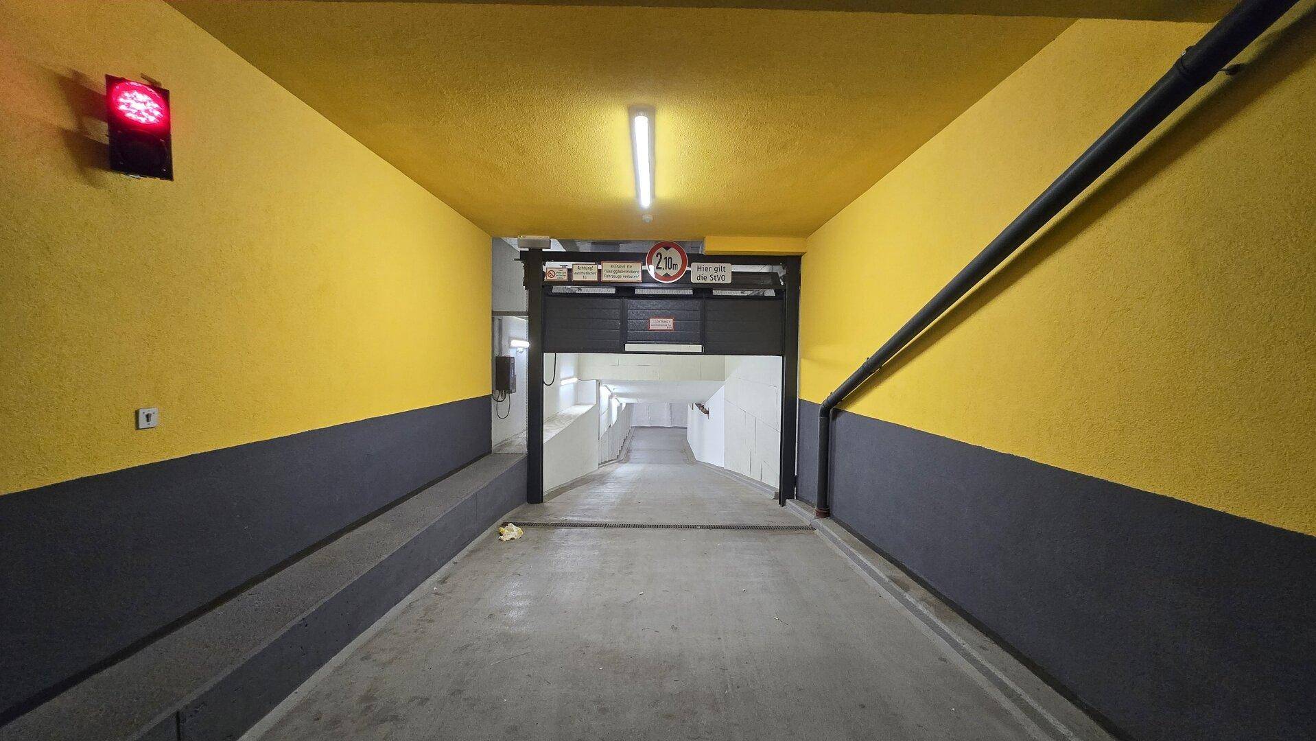 Einfahrt zu einer Tiefgarage mit gelb-grauen Wänden, Betonrampe und einer roten Ampel zur Verkehrsregelung.